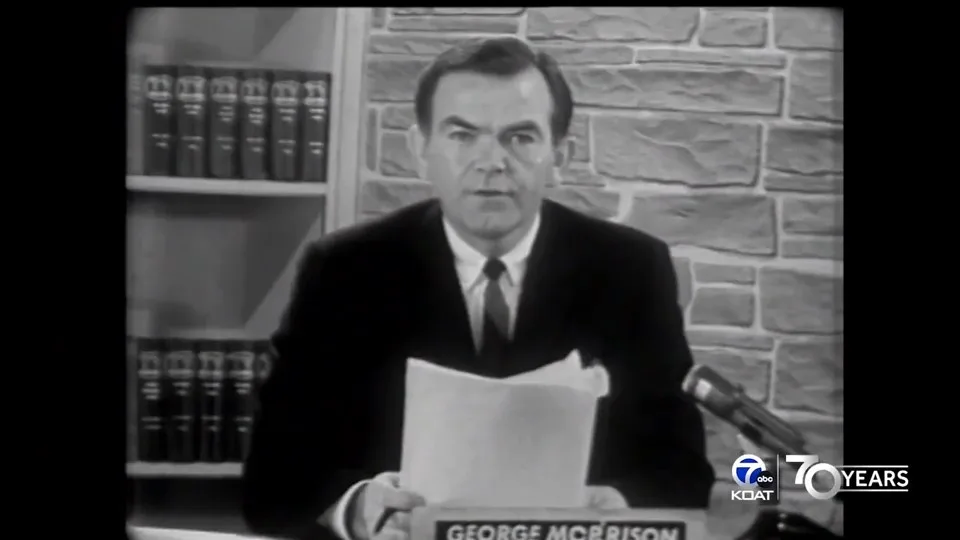 A black-and-white archival photo shows a man in a suit and tie sitting at a desk with a microphone, holding papers. Behind…