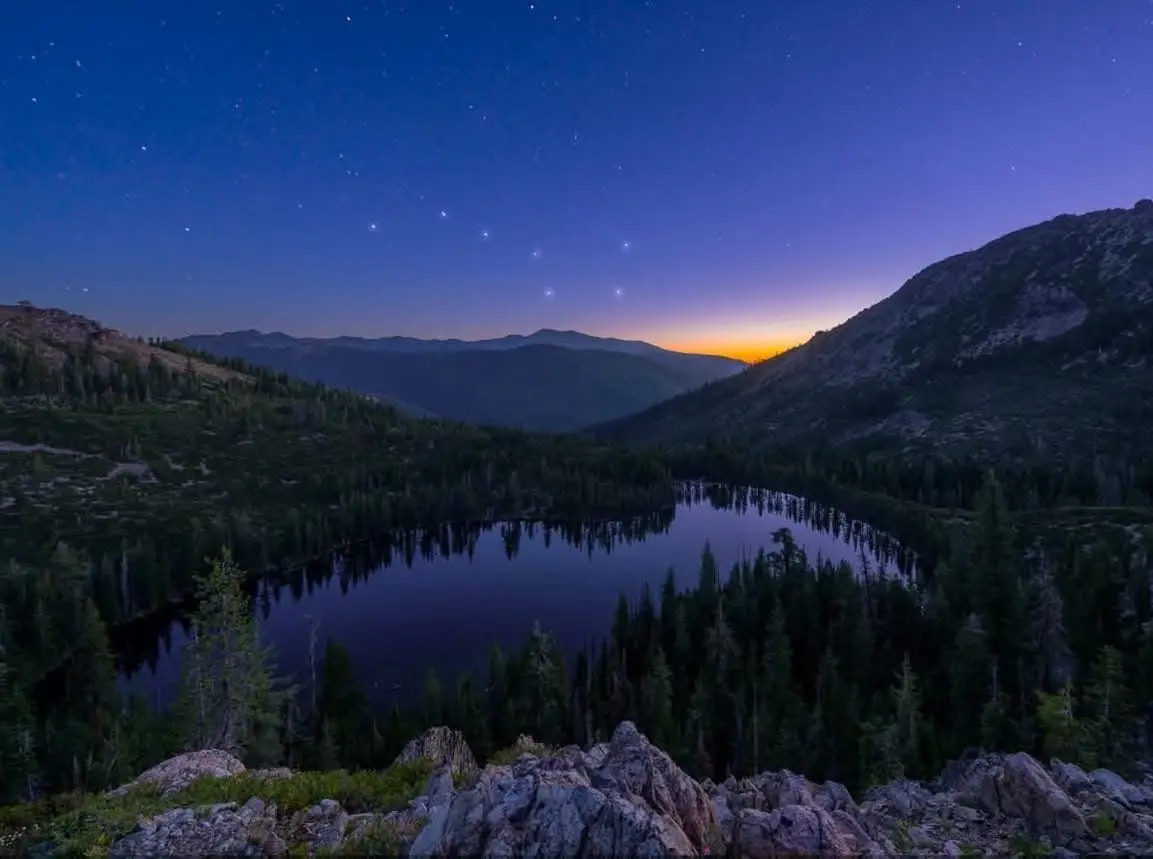 A serene night landscape featuring a calm lake reflecting stars in a valley surrounded by mountains, with a faint orange…