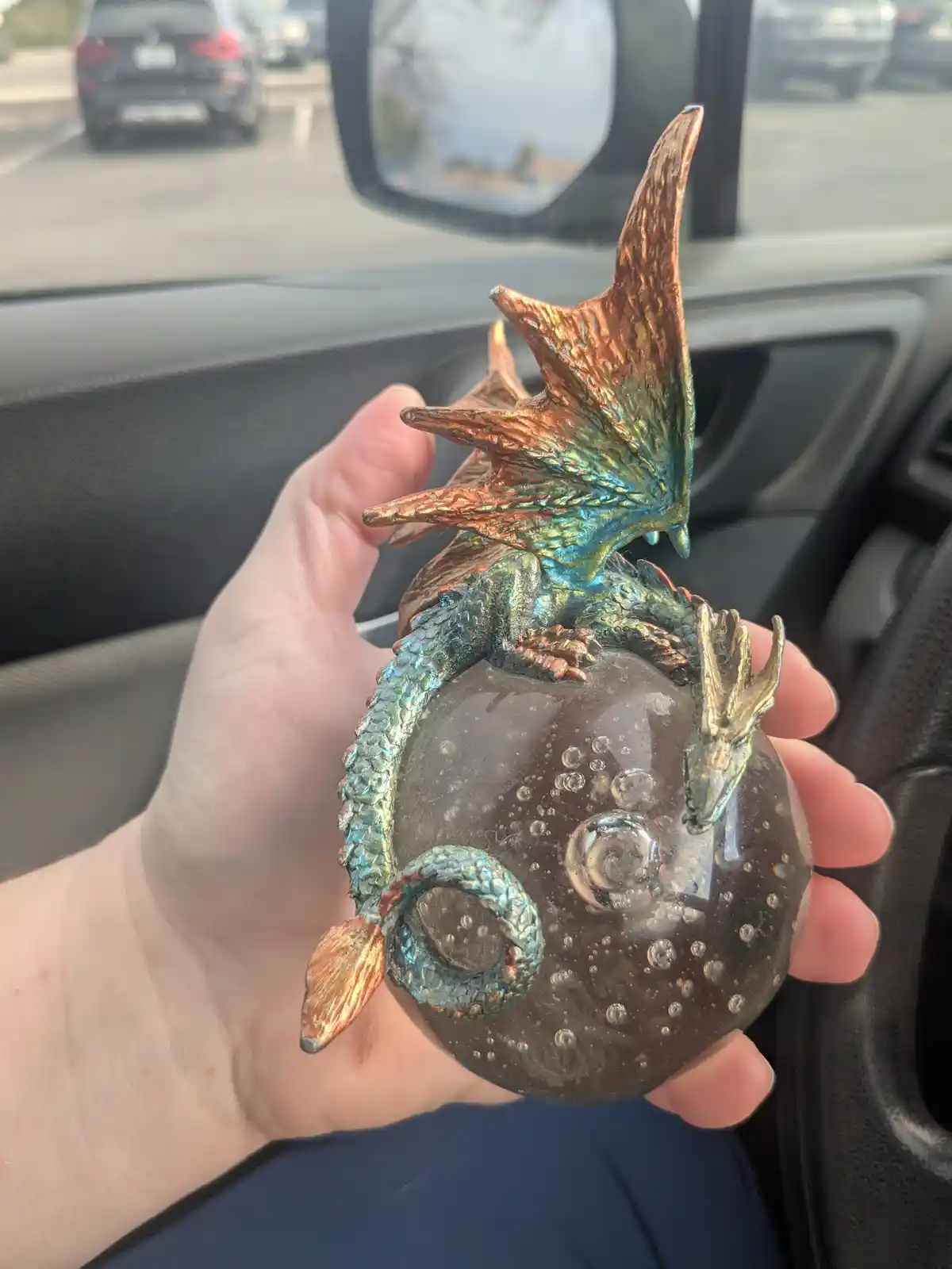 A hand holds a small, iridescent dragon figurine perched atop a clear, bubble-filled orb inside a car. The background shows…