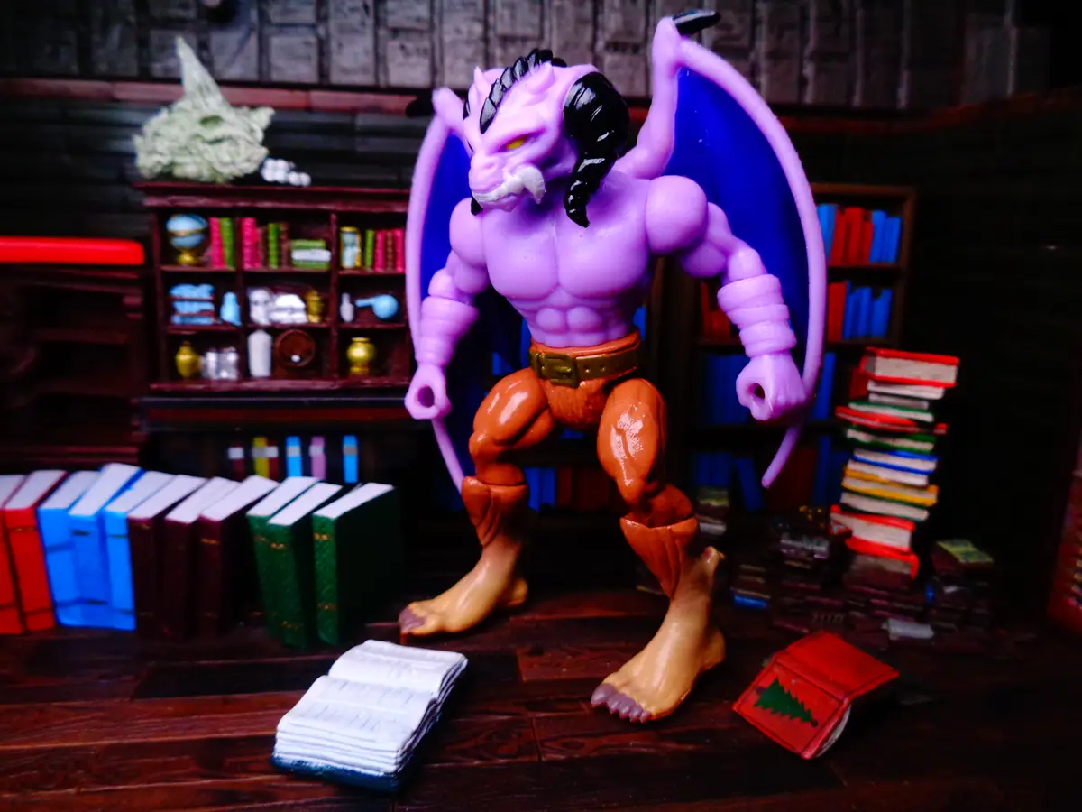 A purple demon toy figure with bat-like wings and horns stands in a miniature library setting surrounded by books. The…