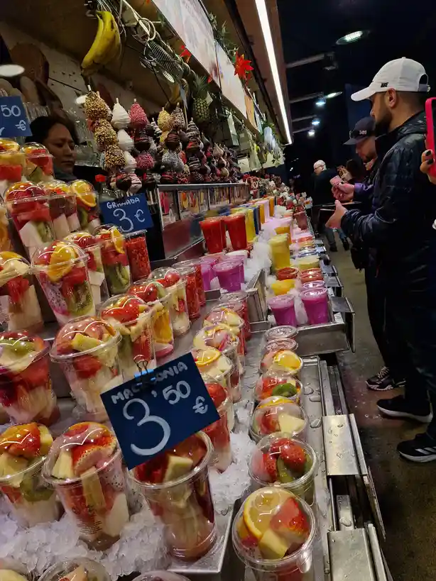 A vibrant market stall displays rows of colorful fruit cups and smoothies on ice, with handwritten price signs showing…