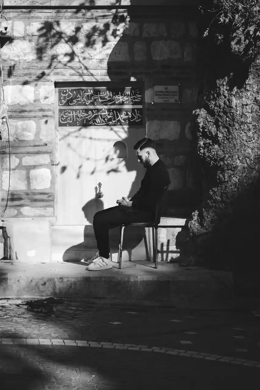 A black-and-white photo shows a man sitting on a chair outside a stone building with Arabic calligraphy above a door. He is…