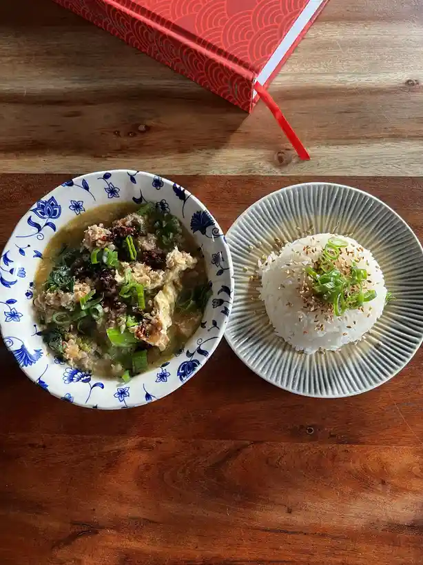 An overhead photo shows two dishes on a wooden table: a bowl of soup with egg, greens, and green onions in a blue floral…