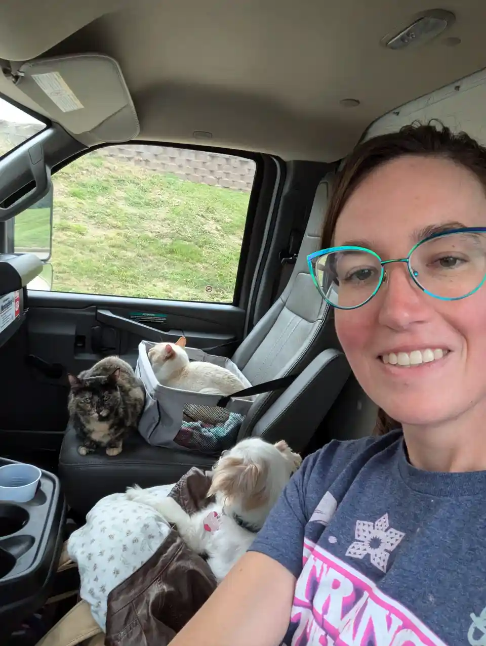 A woman with glasses smiles at the camera from inside a van, surrounded by three pets: a small white dog in her lap, a…