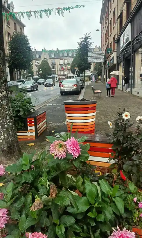 A street scene in a French town with colorful striped planters, flowers, and a road sign pointing to Argentan, Vire…