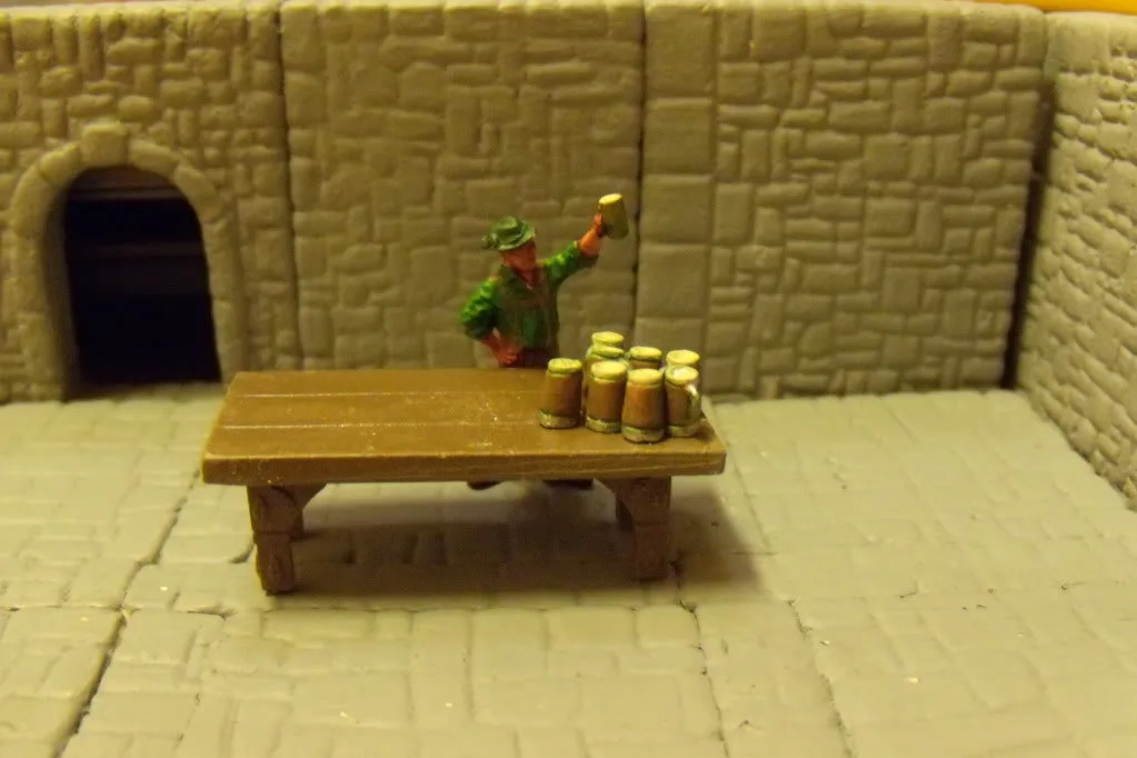 A miniature figurine of a man in a green hat and jacket stands behind a wooden table in a stone-walled diorama, holding up a…