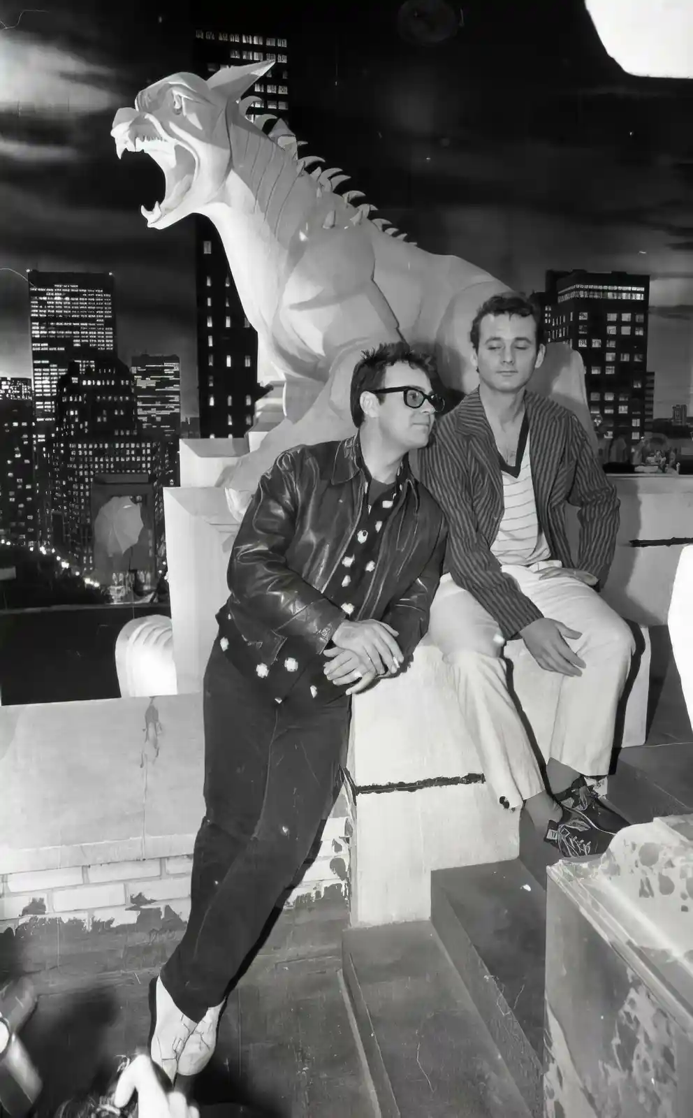 A black-and-white photo shows two men sitting on a rooftop ledge beside a large, roaring dragon statue, with a city skyline…