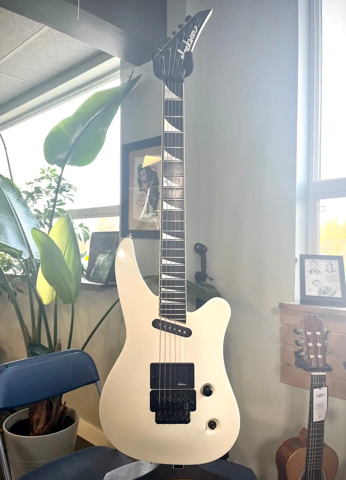 A white Jackson electric guitar with a black fretboard and headstock is standing upright in a room, leaning against a wall…