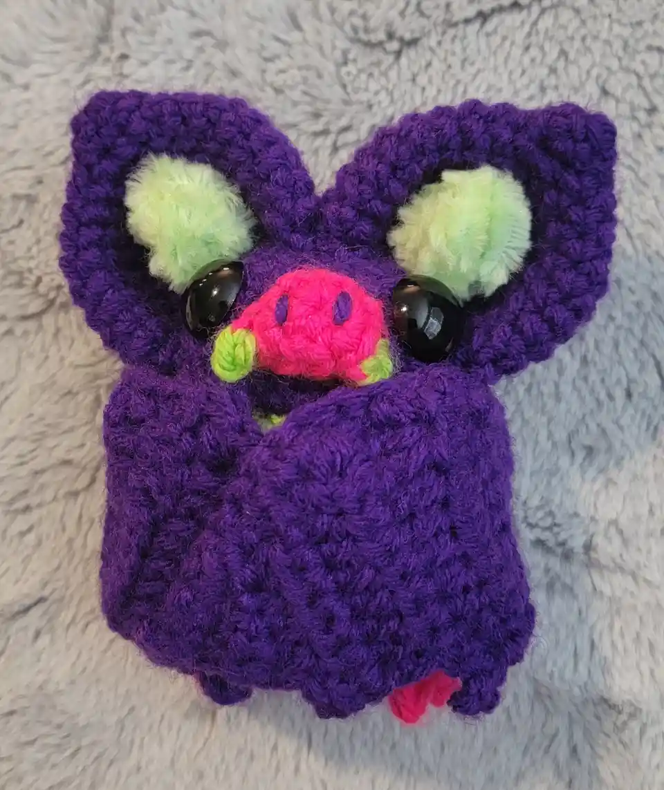 A handmade purple crocheted plush toy resembling a bat or similar creature, featuring large green fuzzy ears, shiny black…