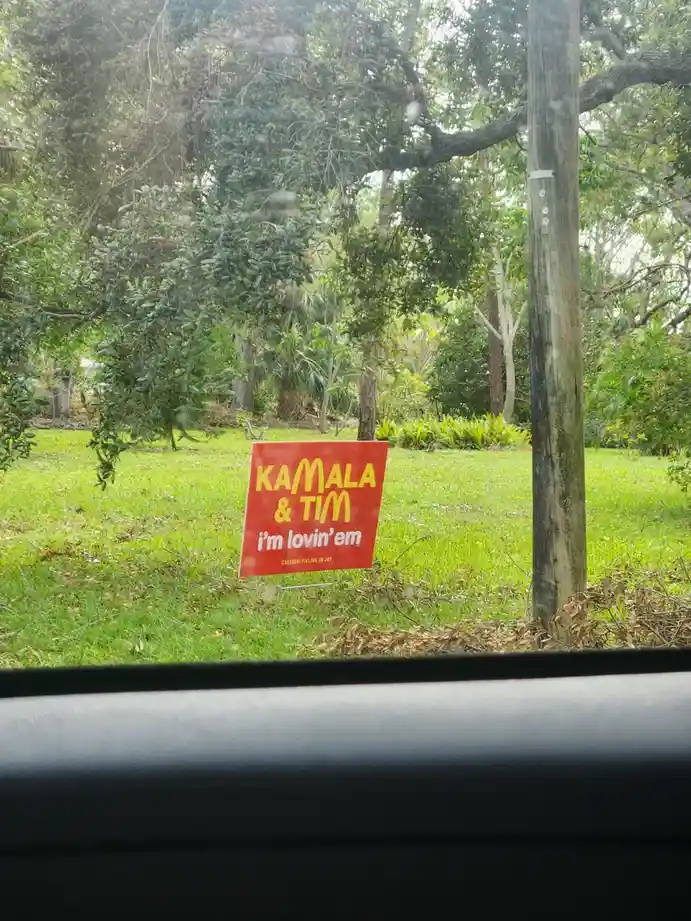 A red yard sign with yellow and white text reading 'KAMALA & TIM' and 'i'm lovin'em' with a small 'CAUTION: FILLING IS JOY'…