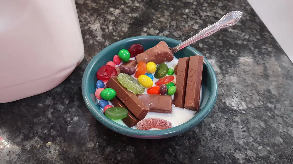 A teal bowl filled with milk and an assortment of candies including Kit Kats, M&Ms, gummy worms, and other assorted sweets…