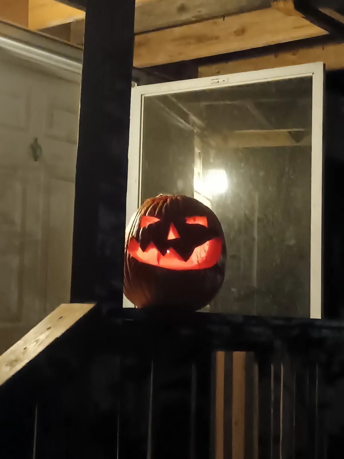 A carved jack-o'-lantern with glowing orange eyes, nose, and mouth sits on a black railing in front of a screen door. The…