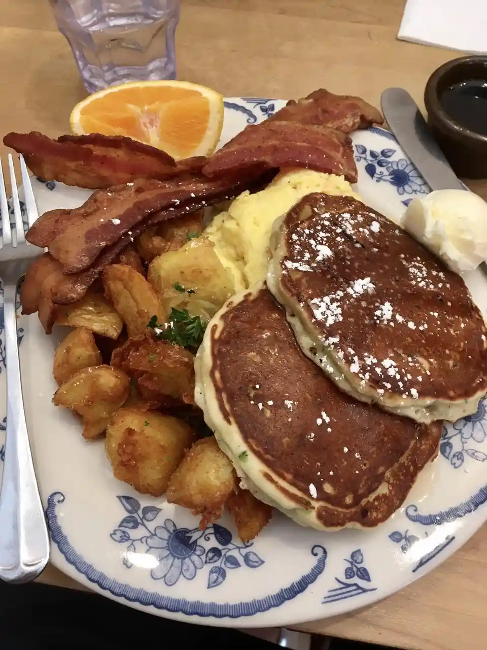 A close-up photo of a hearty breakfast plate featuring two pancakes dusted with powdered sugar, crispy bacon, scrambled…
