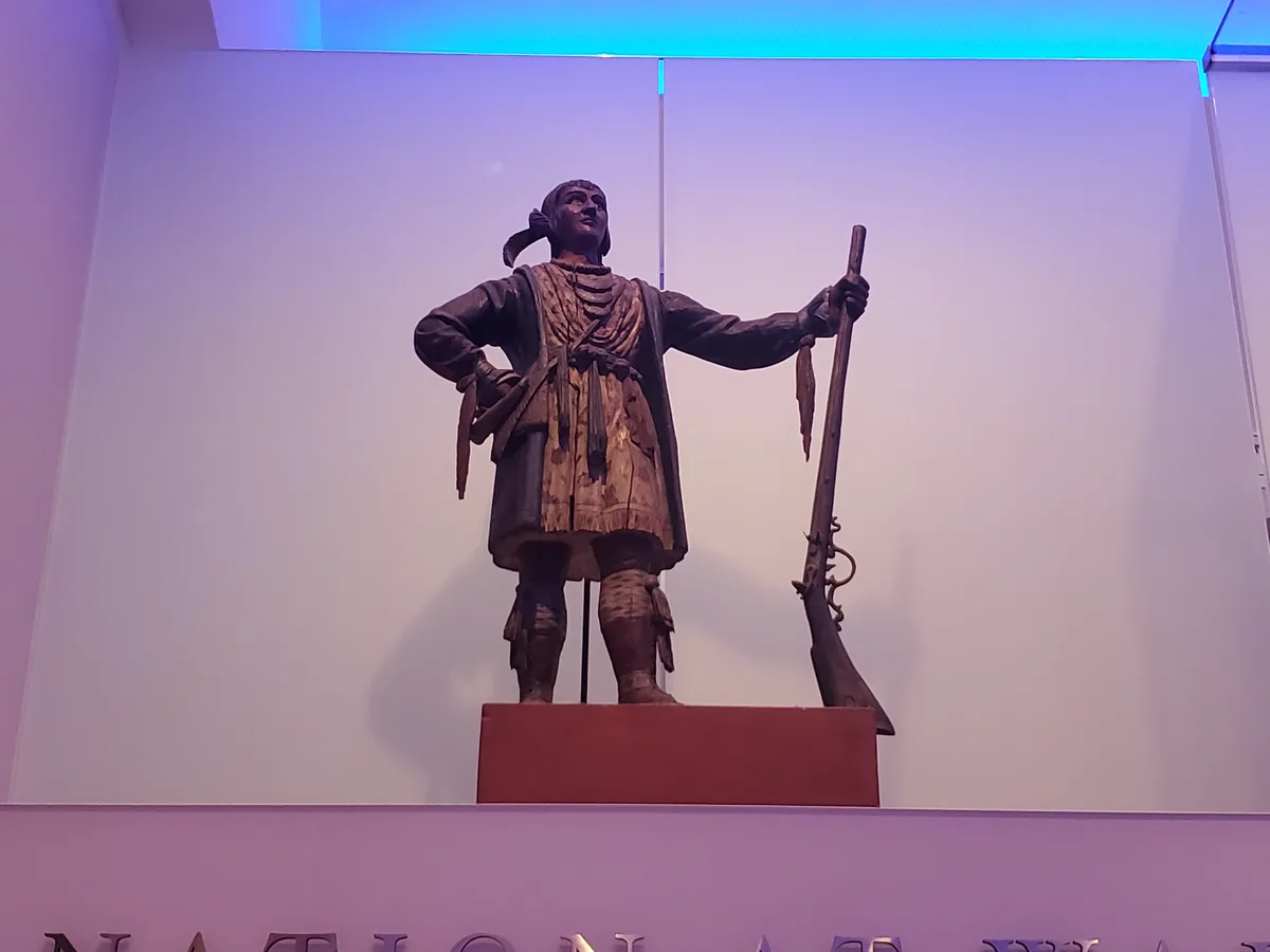 A wooden statue of a person in historical attire holding a musket stands on a pedestal in a museum-like setting under blue…