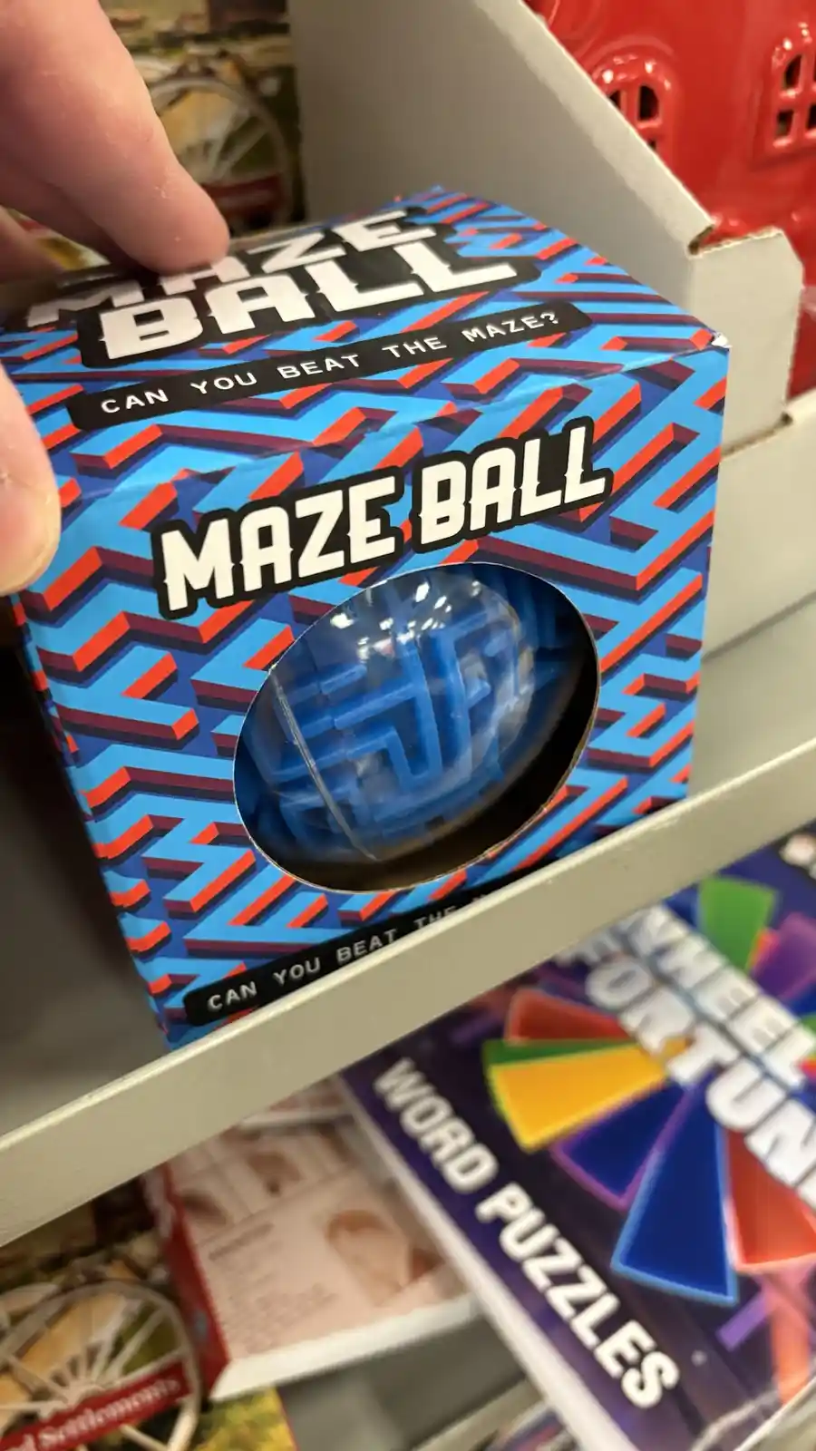 A hand holds a 'Maze Ball' puzzle toy box featuring a blue and red geometric maze pattern, with a circular window revealing…