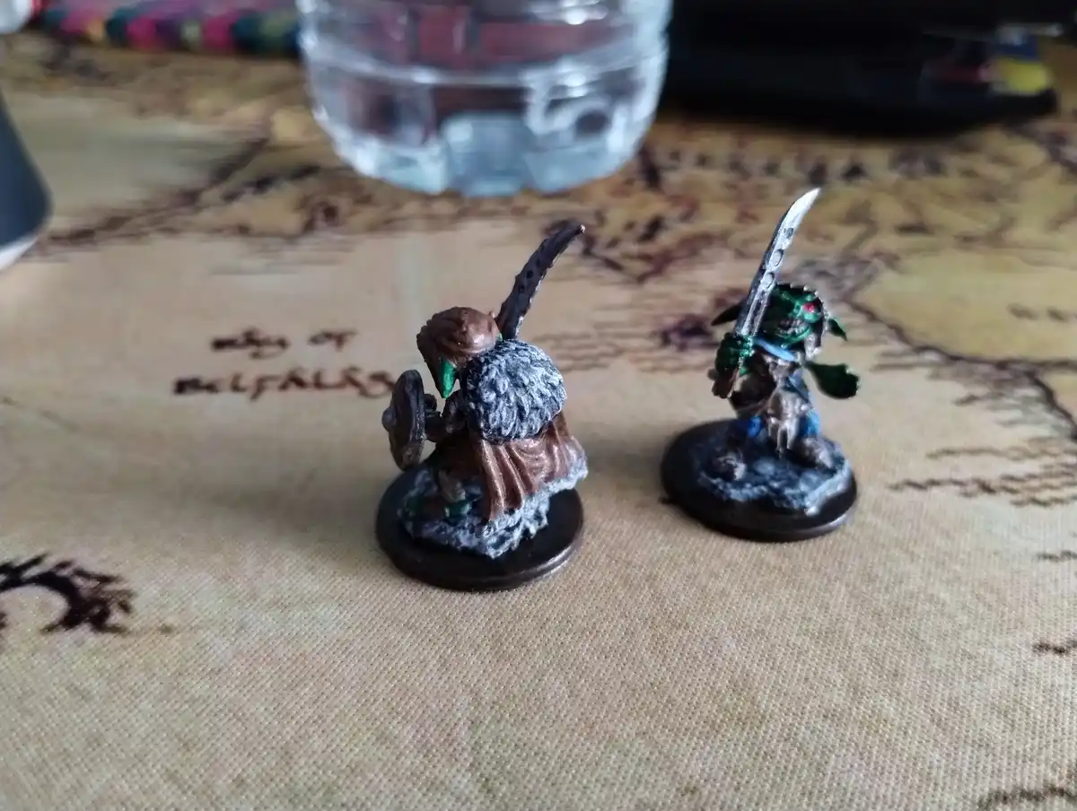 Two painted fantasy miniatures, one holding a shield and curved blade, the other wielding a sword, stand on a textured map…