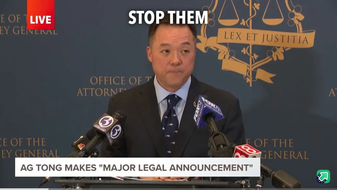 A man in a suit and tie stands at a podium with microphones from news outlets like FOX 3 and CBS 8, in front of a backdrop…