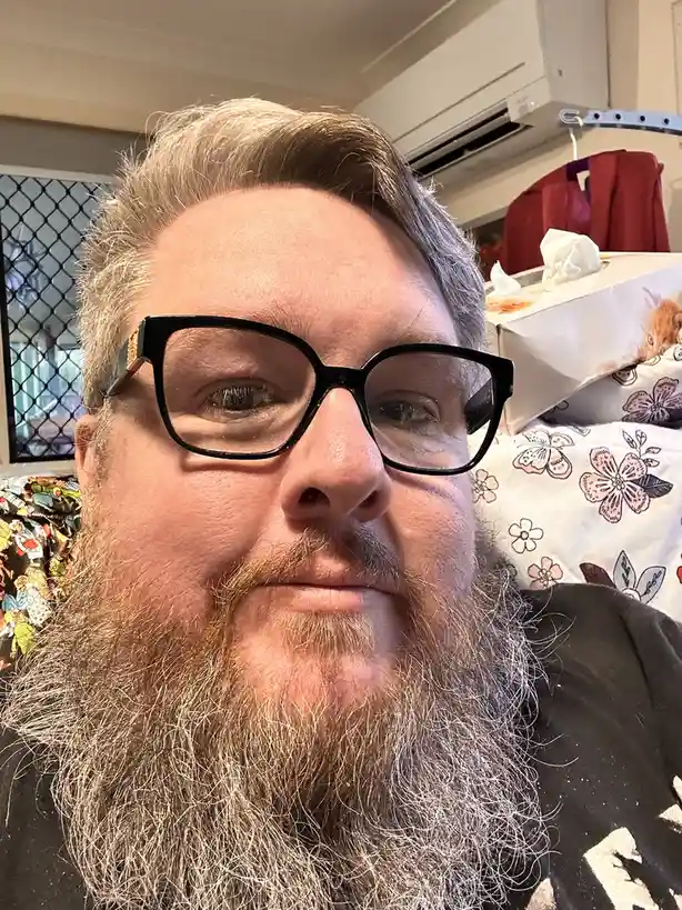 A close-up selfie of a man with a long, graying beard and black-rimmed glasses, looking directly at the camera with a…