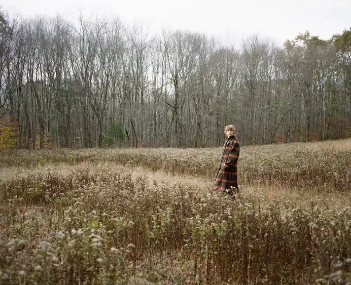 A person with short, light-colored hair stands alone in a field of tall, dry, brownish vegetation under an overcast sky…