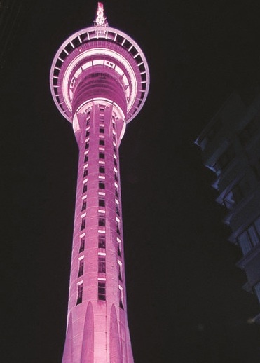 A tall, slender tower illuminated in pink light against a dark night sky, with a circular observation deck near the top and…
