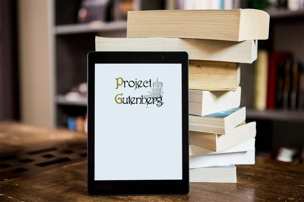 An e-reader displaying the 'Project Gutenberg' logo sits on a wooden table next to a tall stack of physical books, with a…