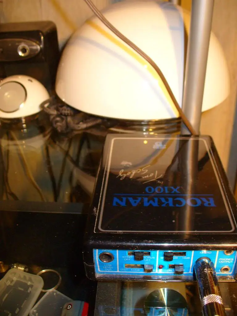 A close-up photo of a black Rockman X100 guitar effects pedal with a handwritten signature reading 'Tom Scholz' on its top…