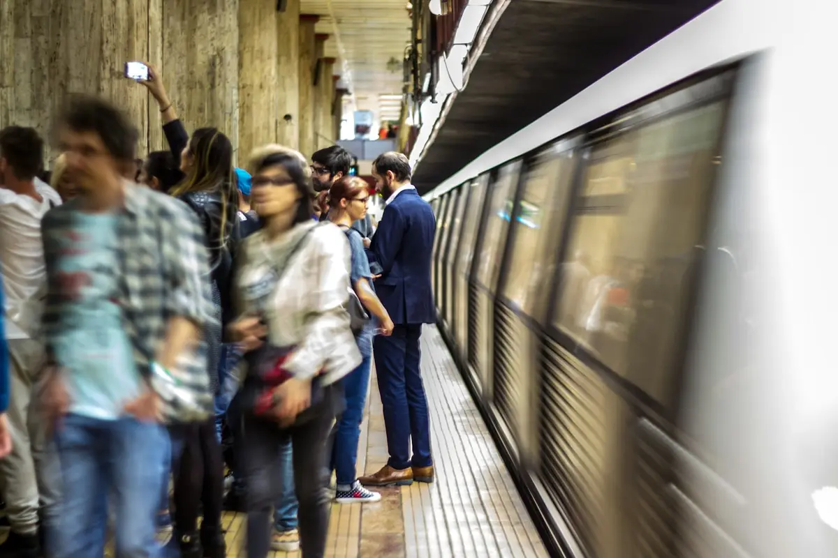 A motion-blurred photograph of people standing on a subway platform as a train speeds past. One person in the background…