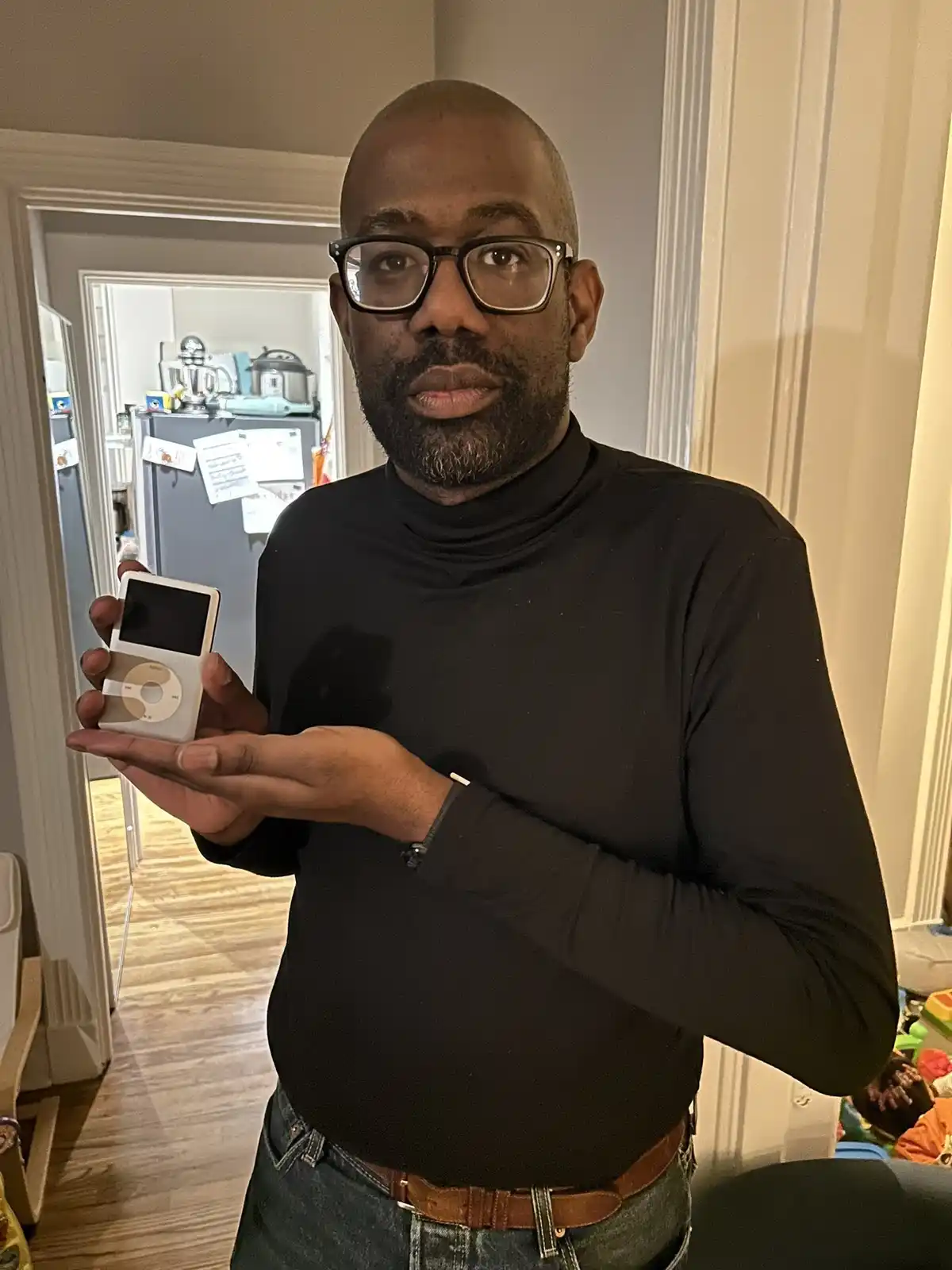 A man with glasses and a black turtleneck holds up a white iPod Classic, looking directly at the camera with a deadpan…