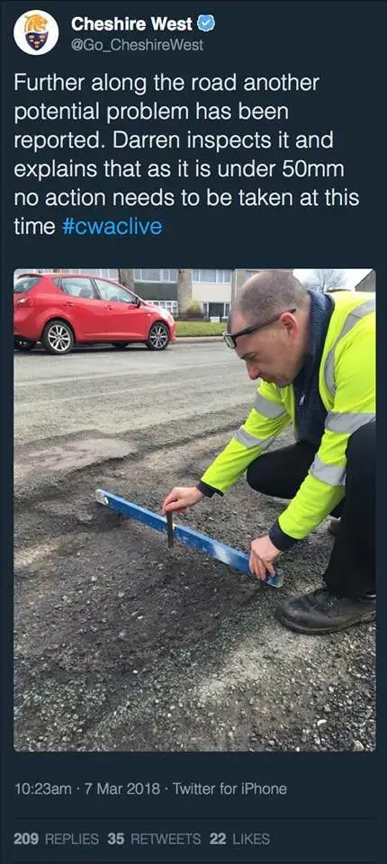 A man in a bright yellow high-visibility jacket and glasses is crouching on a road, using a blue spirit level to measure a…