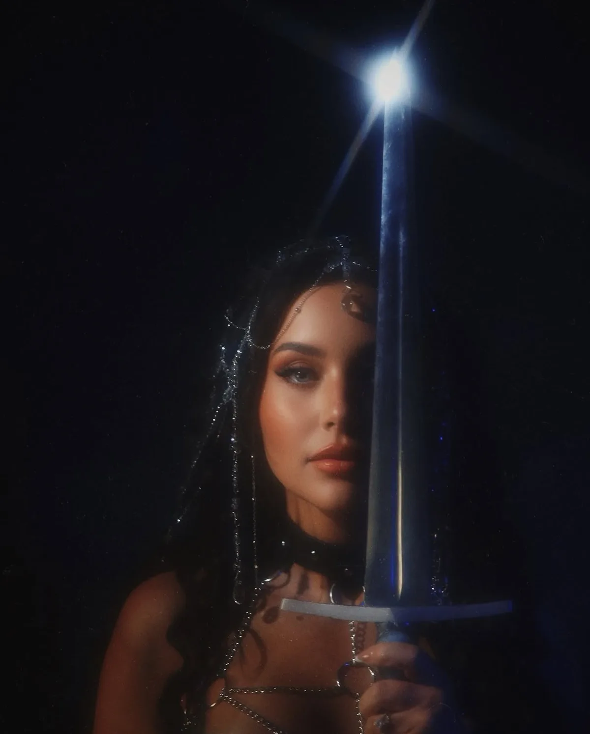 A woman with dark hair and ornate silver chains in her hair and on her body holds a sword vertically, its tip glowing…