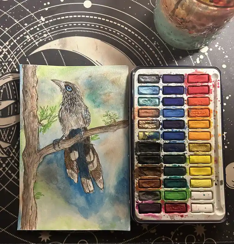 A watercolor painting of a bird with blue eyes perched on a branch next to a heavily used watercolor palette, with the…