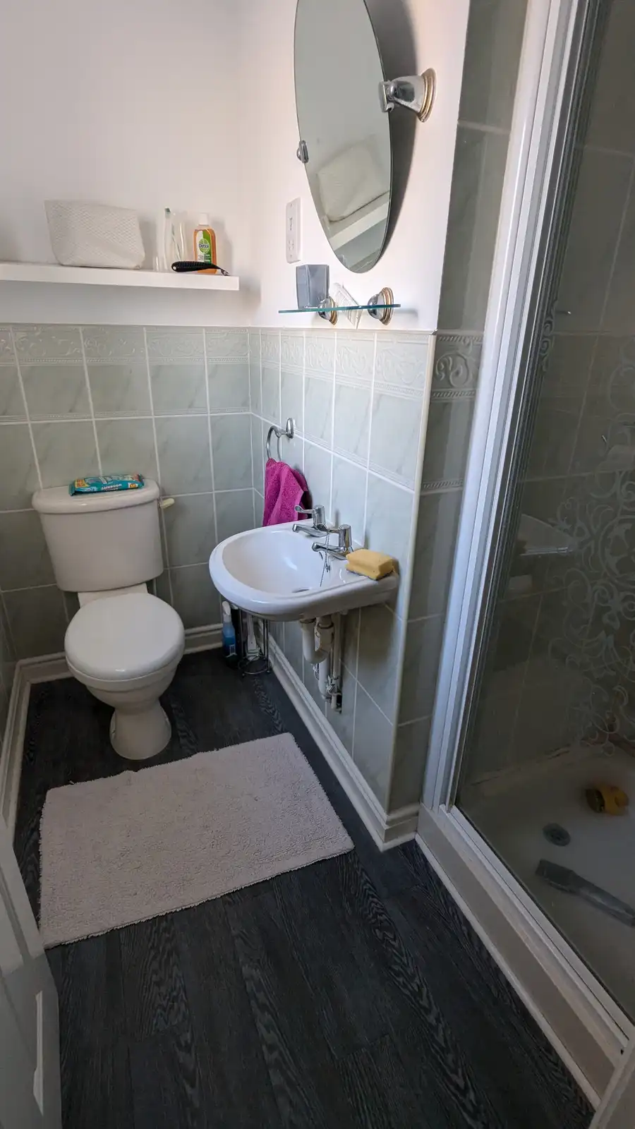A small bathroom with a toilet, sink, and shower stall, featuring light green tiled walls, dark wood-style flooring, and a…