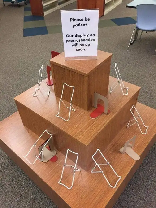 A tiered wooden display stand in a library or office setting, adorned with numerous empty, white wire display easels. A sign…