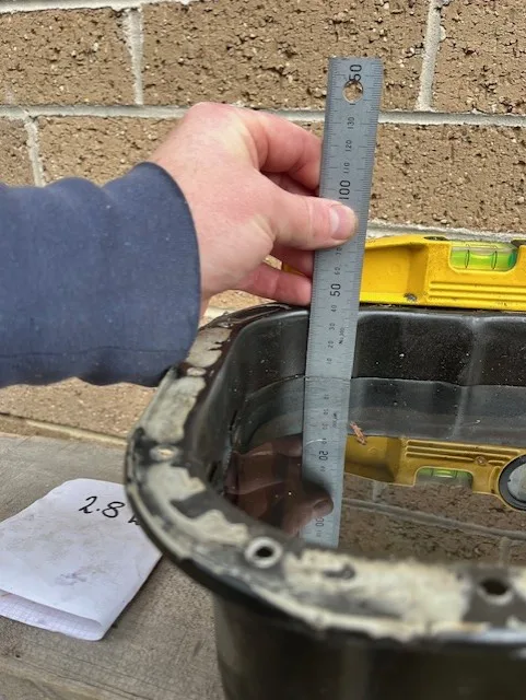 A hand holds a metal ruler vertically inside a black plastic tub filled with water, measuring the water level. A yellow…