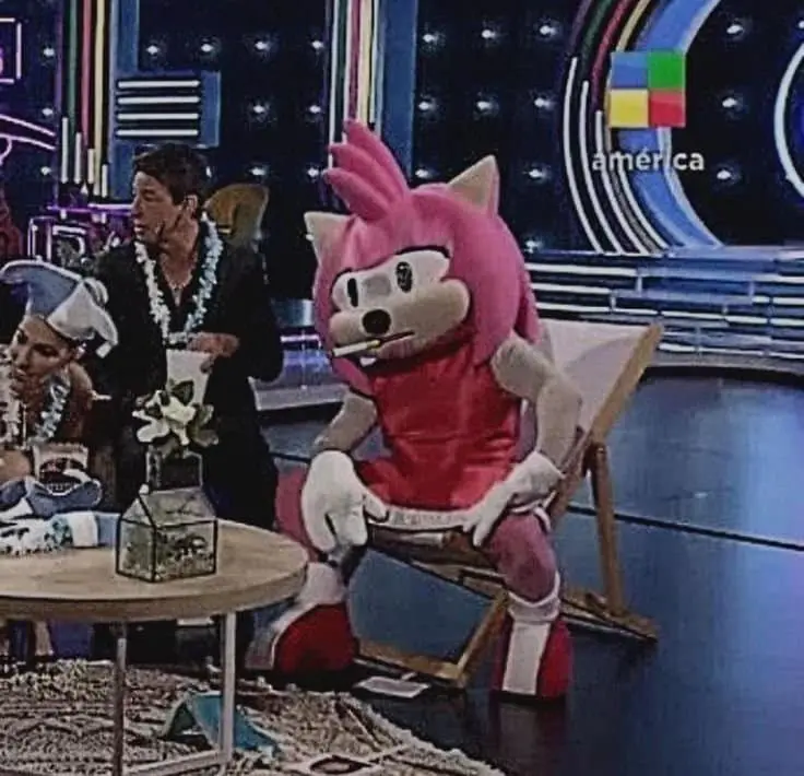 A person in a pink hedgehog mascot costume sits in a beach chair on a TV show set, holding a cigarette. Behind them, a man…