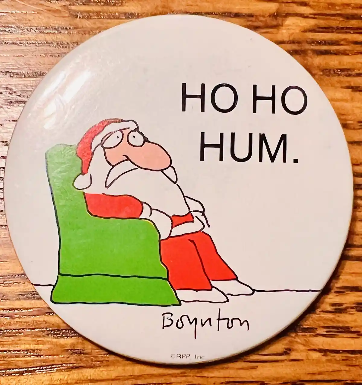 A round white button features a cartoon Santa Claus sitting in a green armchair, looking bored or unenthusiastic. The text…