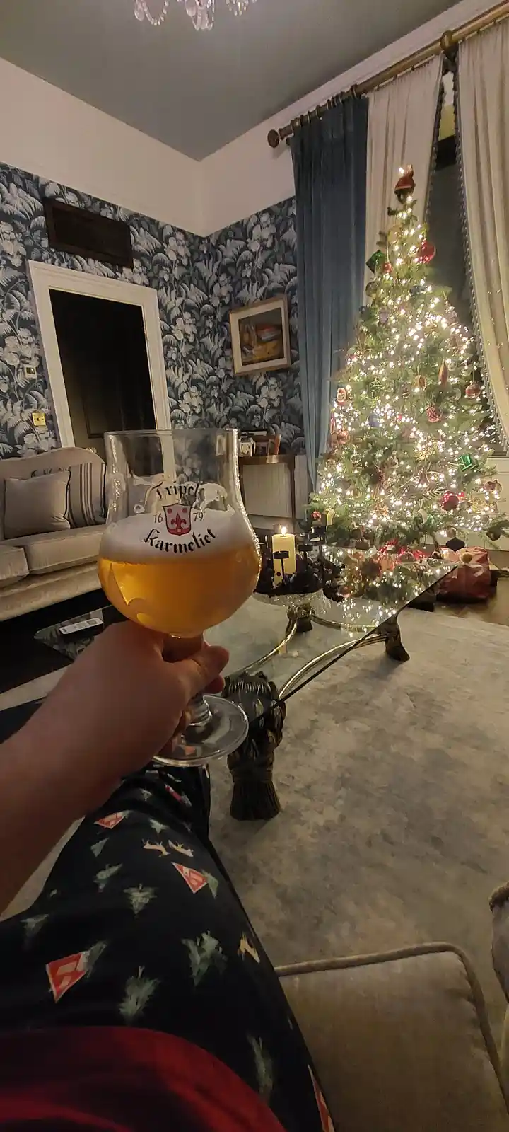 A person holds a glass of Tripel Karmeliet beer in a cozy living room with a lit Christmas tree in the background. The room…