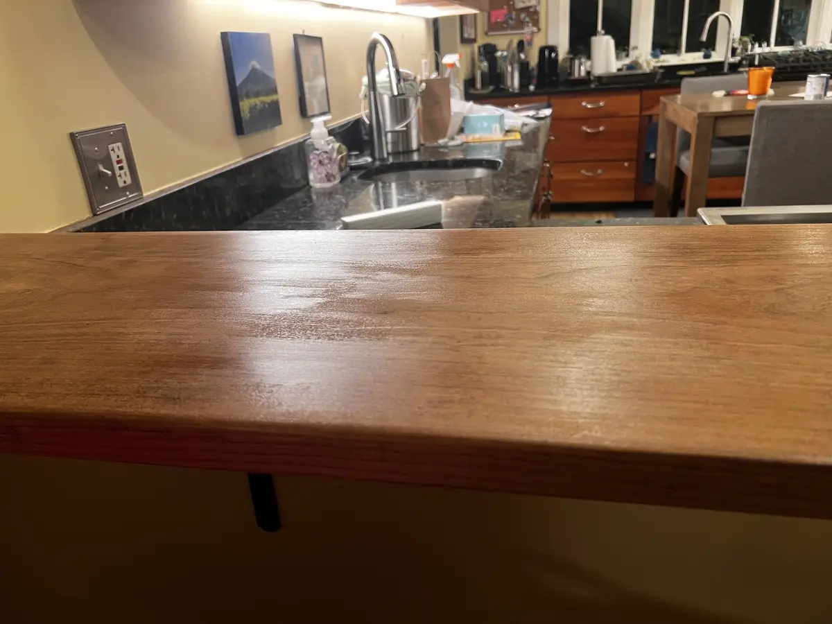 A low-angle photo of a clean, polished wooden kitchen counter in the foreground, with a dark granite countertop and sink…