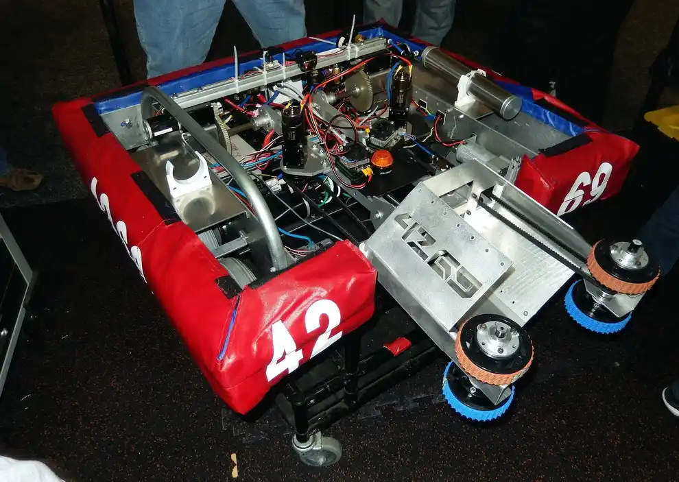 A red and silver robot combat machine with exposed wiring, gears, and wheels, marked with the number 42 on a padded side…