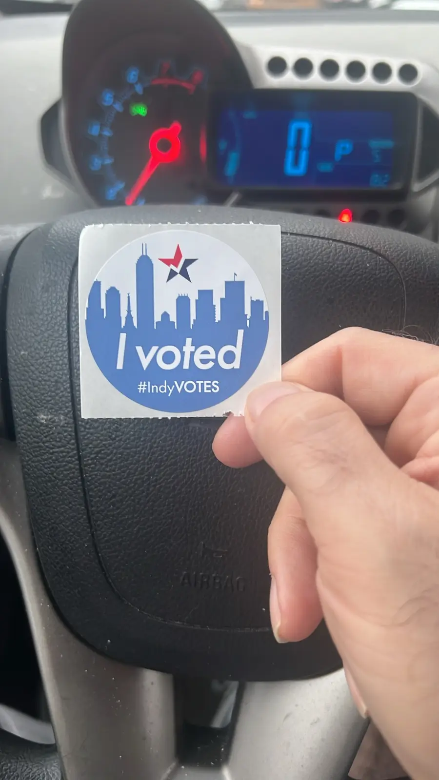 A hand holds an 'I voted #IndyVOTES' sticker over a car's steering wheel, with the dashboard visible in the background…