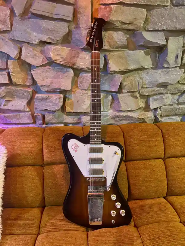 A Gibson electric guitar with a white pickguard and sunburst finish is propped upright on a textured orange couch, set…