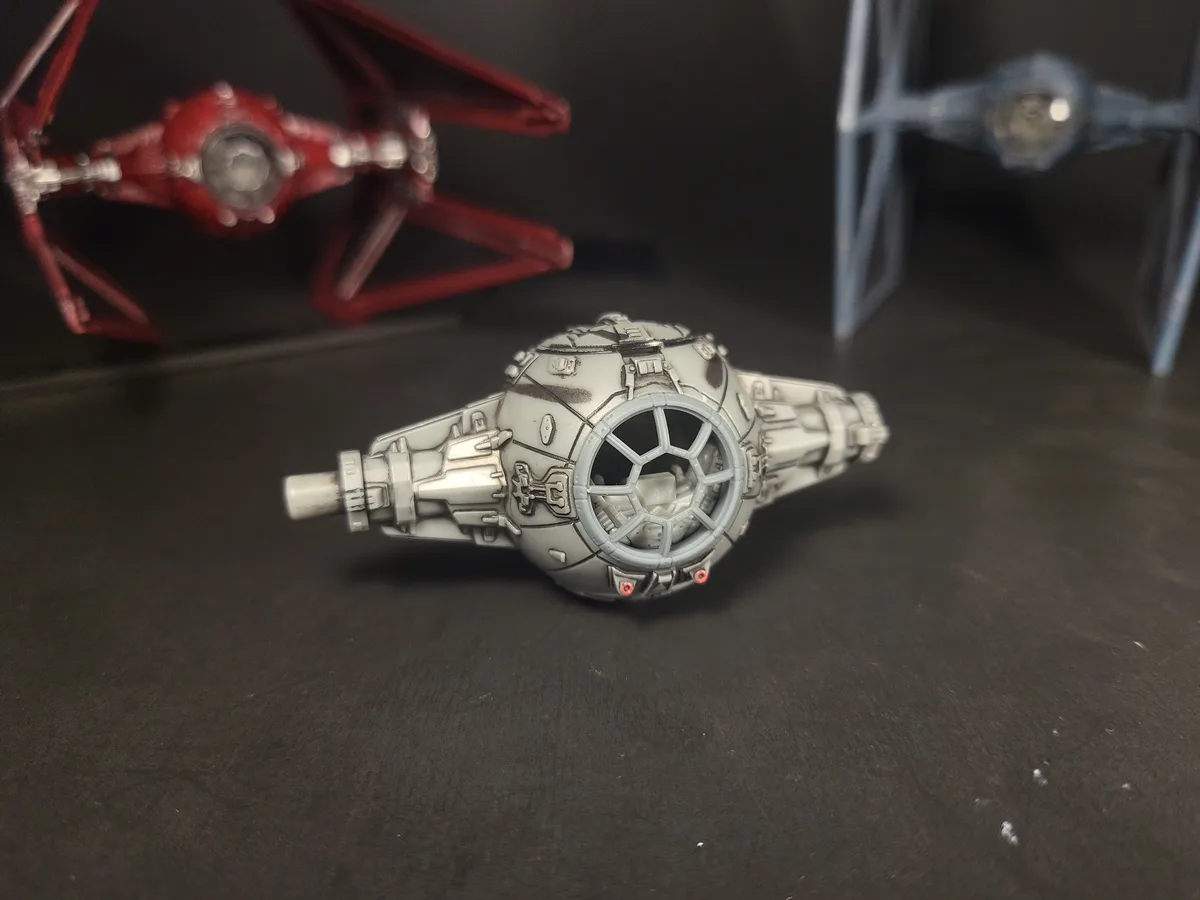 A close-up photo shows a small, gray, spherical Star Wars TIE fighter model lying on its side on a dark surface. In the…