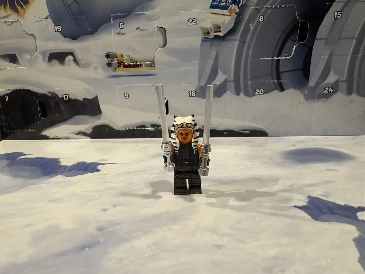 A LEGO minifigure of Ahsoka Tano, holding two lightsabers, stands in front of a LEGO Star Wars Advent calendar with…