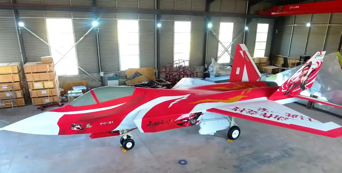 A red and white fighter jet, identified as FC-31, is parked inside a hangar. The aircraft features cartoon characters and…