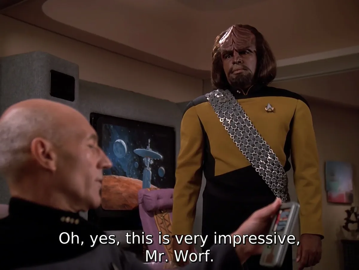 A still from Star Trek: The Next Generation shows Captain Picard in profile, holding a device, while Worf stands behind him…