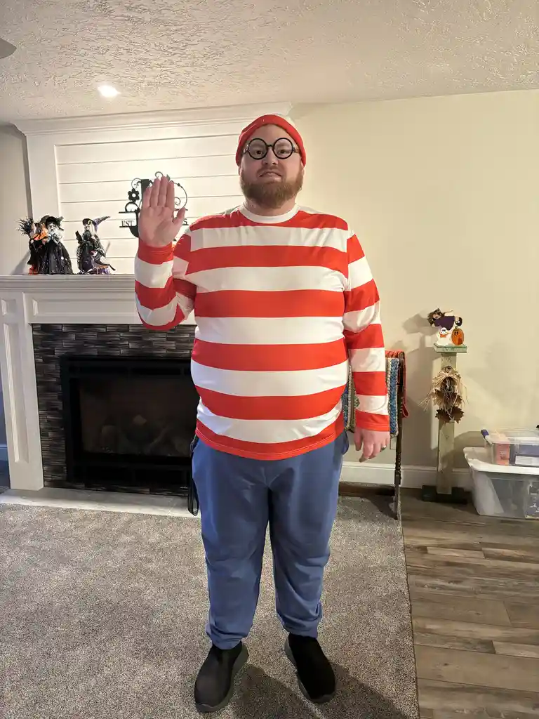 A man with a beard, wearing a red and white striped shirt, round black glasses, and a red beanie, stands indoors in front of…