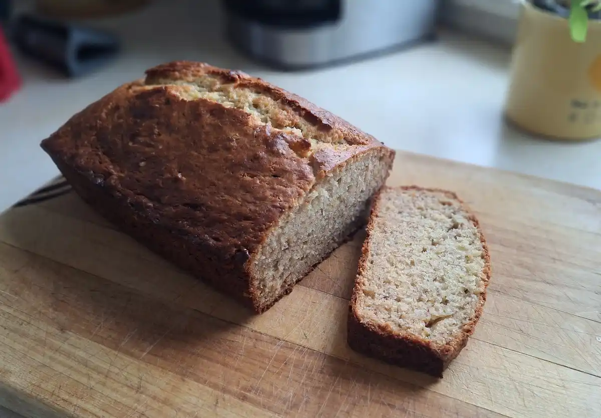 A freshly baked loaf of banana bread with one slice cut and placed beside it on a well-used wooden cutting board. The bread…