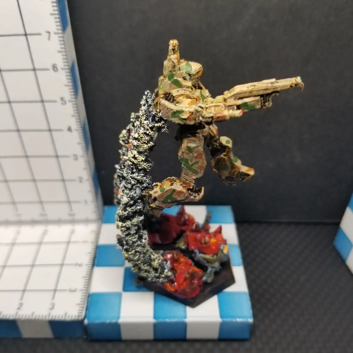 A heavily weathered and camouflaged miniature soldier figurine, posed mid-action with a rifle, stands on a hexagonal base…