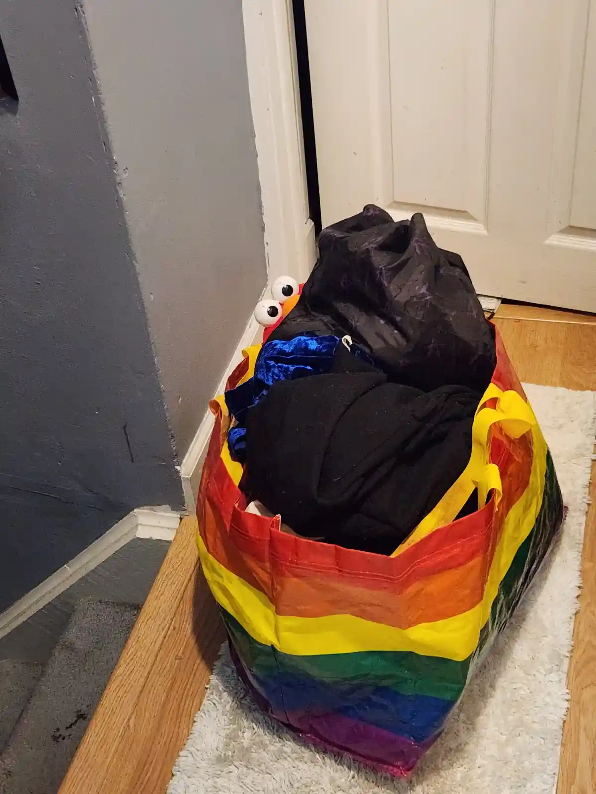 A rainbow-colored laundry bag filled with dark clothes sits on a white rug near a door, with two large googly eyes and an…