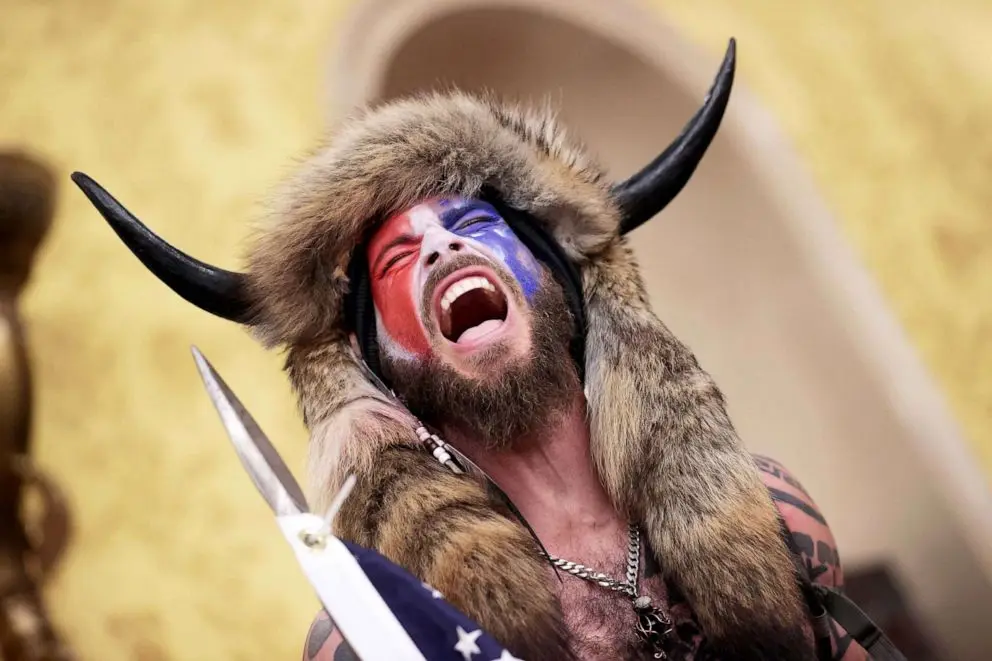 A man with a fur hat featuring black horns and face paint in red, white, and blue is screaming with his mouth wide open. He…